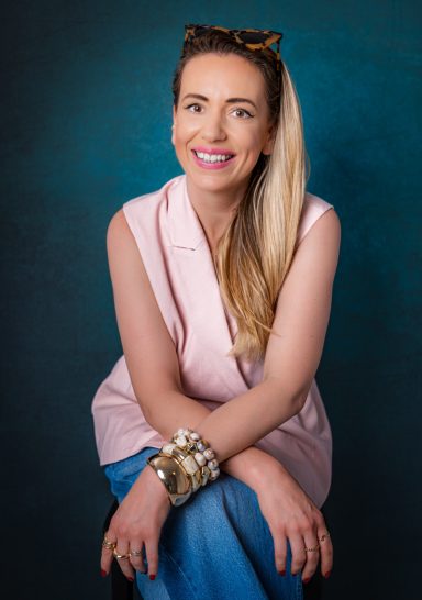 Smiling blonde woman in a light pink blazer leaning forward during a studio portrait session.