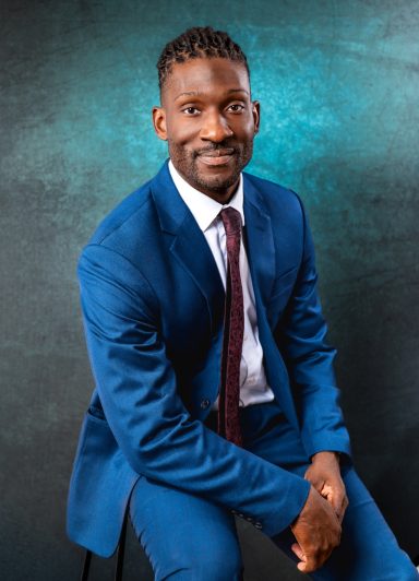 Seated professional portrait of a man in a blue suit and tie against a textured teal studio background.