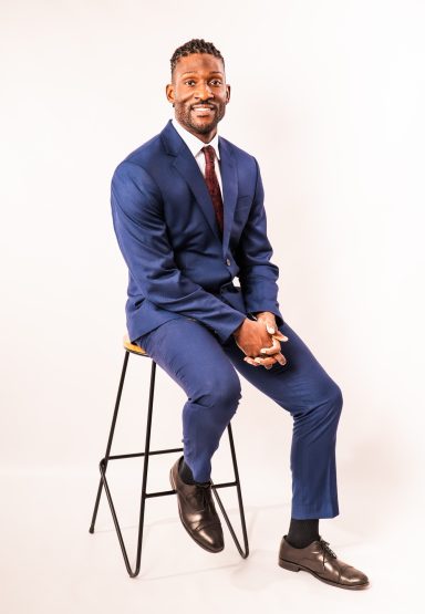Professional business portrait of a smiling man in a blue suit and burgundy tie seated on a studio stool.