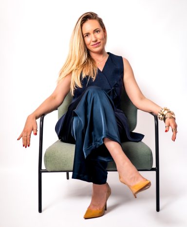 Full-body portrait of a smiling blonde woman seated on a chair with her legs crossed, wearing a navy outfit.