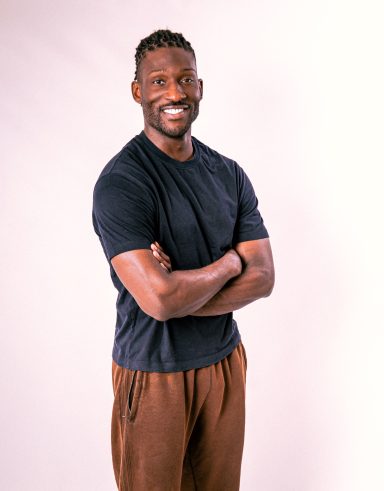 Smiling man wearing a dark T-shirt, standing with his arms crossed against a light studio background.