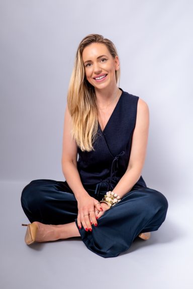 Relaxed seated portrait of a smiling blonde woman wearing a navy sleeveless outfit against a light background.