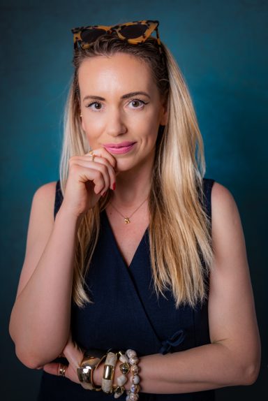 Studio portrait of a blonde woman in a navy sleeveless top with her hand near her chin against a blue background.