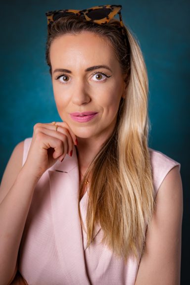 Close-up studio headshot of a blonde woman wearing a light pink blazer against a blue background.